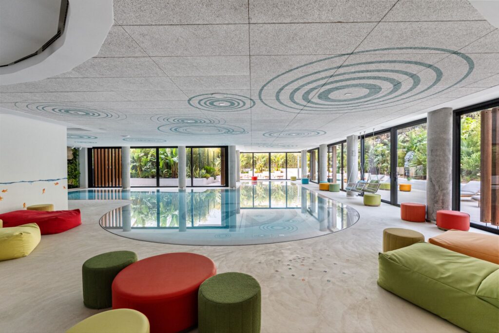 Immeuble d'appartements de luxe avec piscine intérieure et coin salon à Playa d'en Bossa, Ibiza, avec des sièges colorés, des baies vitrées et une vue extérieure tropicale.