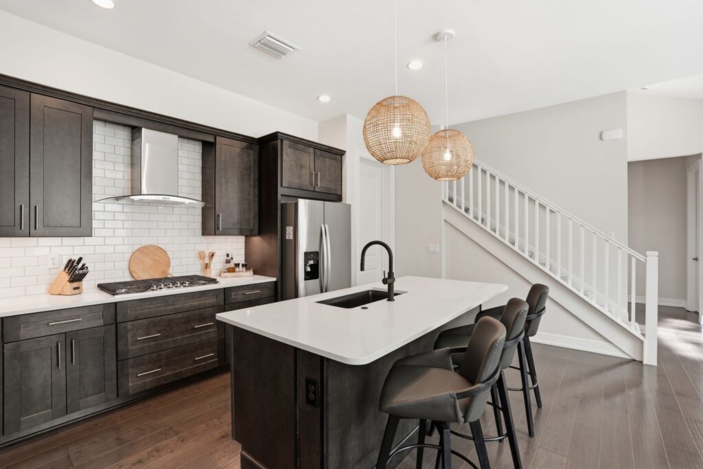Cuisine moderne à aire ouverte comprenant des armoires en bois sombre, un îlot en quartz blanc avec des sièges, un dosseret en carrelage métro blanc et une vue sur l'escalier au 9194 Cayman Drive à Naples, en Floride.